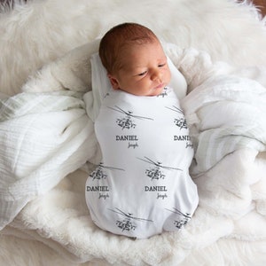 May include: A newborn baby swaddled in a white swaddle blanket with a black helicopter and the name "Daniel Joseph" repeated on it. The baby is lying on a white fluffy rug.