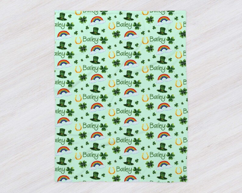 May include: A green and white baby blanket with a repeating pattern of shamrocks, rainbows, leprechaun hats, and gold horseshoes. The name "Bailey" is repeated throughout the pattern.