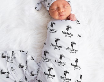 May include: A white swaddle blanket and matching hat with a black and white pattern of the text "Hudson Samuel" and a graphic of a power line. The baby is sleeping peacefully in the swaddle.