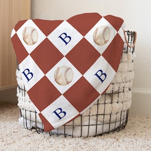 May include: A red and white checkered throw blanket with baseballs and the letter "B" printed on it. The blanket is folded and placed in a wire basket.