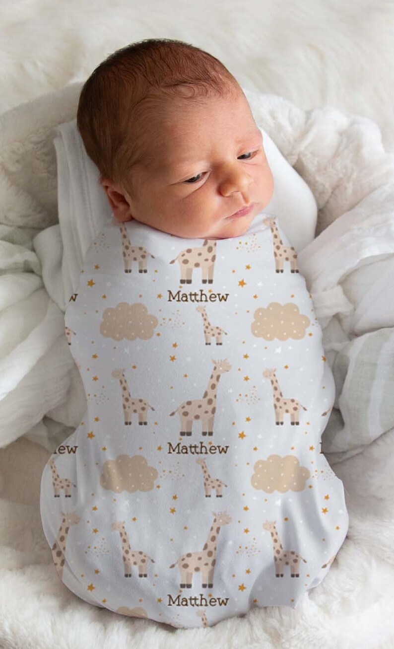 May include: A white swaddle blanket with a pattern of brown giraffes, gold stars, and beige clouds. The name "Matthew" is printed three times on the blanket.