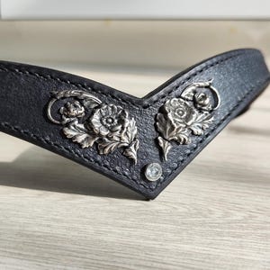 May include: Black leather headband with silver floral detailing. The headband has two ornate floral designs and a small, clear gem. The leather is stitched along the edges.