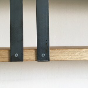 May include: Two black metal brackets are attached to a light brown wooden shelf. The brackets are secured with screws.