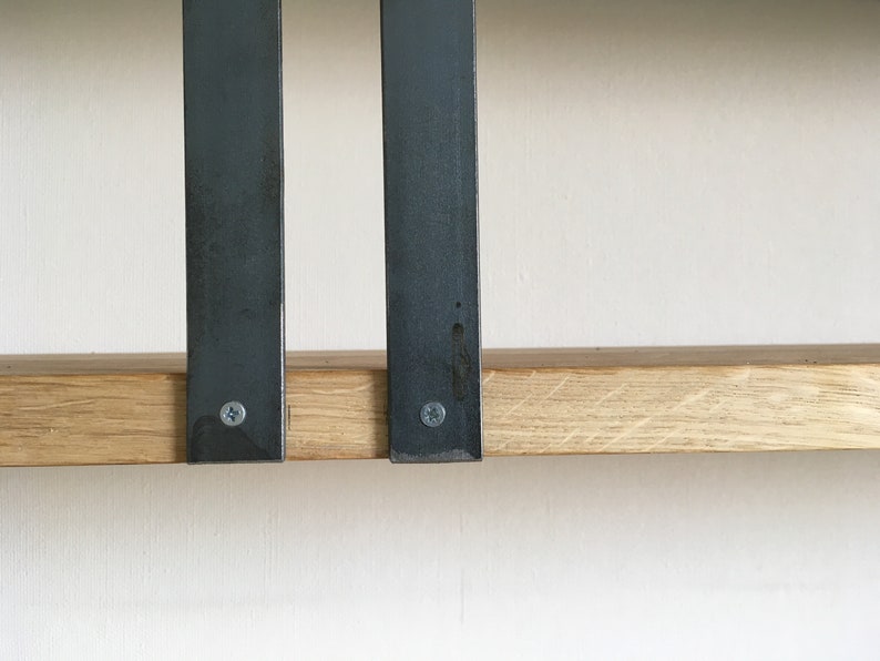 May include: Two black metal brackets are attached to a light brown wooden shelf. The brackets are secured with screws.