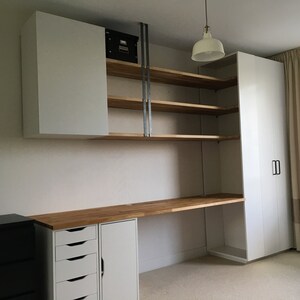 May include: A white desk with a wood top and drawers. The desk is against a white wall with white cabinets and shelves above it. There is a white closet with a door on the right side of the image.