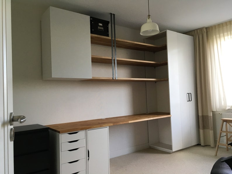 May include: A white desk with a wood top and drawers. The desk is against a white wall with white cabinets and shelves above it. There is a white closet with a door on the right side of the image.