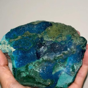 May include: A raw mineral specimen featuring a vibrant mix of deep blue, turquoise, and forest green hues. The surface displays a crystalline, textured appearance with varied geological patterns, showing natural color transitions across the stone.