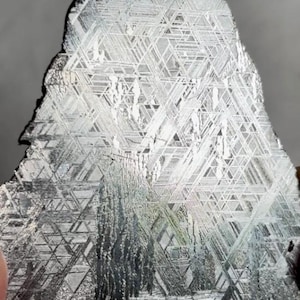 May include: A polished metallic meteorite slice featuring a distinct Widmanstätten pattern of intersecting crystalline lines. The surface displays a silver and grey color palette with a reflective, geometric grid structure across the entire face.
