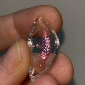 May include: A clear, teardrop-shaped gemstone with internal iridescent pink and purple inclusions. The polished stone has a slightly textured surface and is held between fingers. Tiny black specks are scattered throughout the gem.