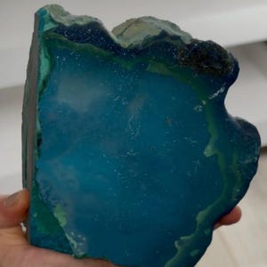 May include: A vibrant, polished agate stone slice, showcasing a deep teal center with a contrasting green and black outer edge. The natural gemstone has a unique, organic shape, perfect for display or crafting.