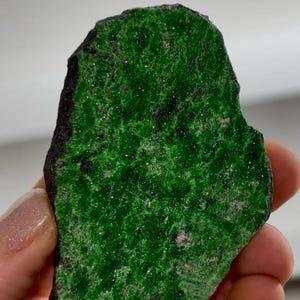 May include: A vibrant green mineral specimen with a rough, dark-colored edge. The surface of the stone is a rich, textured green, with small, sparkling inclusions. The shape is irregular, showcasing its natural form. A unique piece for collectors.