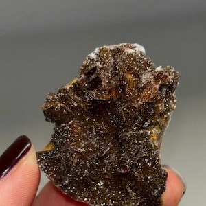 May include: A dark brown mineral specimen with a rough, textured surface and small, sparkling crystals. The mineral has a layered appearance with areas of lighter color. The image shows the mineral held in a hand.