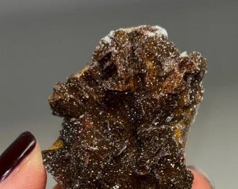 Puede incluir: Un espécimen mineral de color marrón oscuro con una superficie rugosa y texturizada y pequeños cristales brillantes. El mineral tiene una apariencia en capas con áreas de color más claro. La imagen muestra el mineral en una mano.
