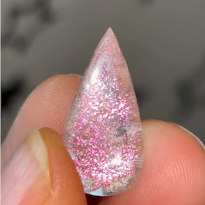 May include: A teardrop-shaped cabochon with a clear, transparent body containing a dense cluster of vibrant, reflective pink glitter particles that catch the light, creating a sparkling effect throughout the interior of the smooth, polished stone.