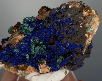 May include: A natural azurite and malachite mineral specimen. The stone features a deep blue color with patches of green and brown, mounted on a clear acrylic stand. The mineral's surface has a rough, textured appearance.
