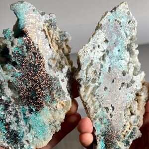 Puede incluir: Dos piezas de mineral en bruto, cada una con una combinación única de colores y texturas. El mineral presenta áreas de color turquesa, negro y cobre, con una superficie rugosa y porosa. Las piezas se sostienen en las manos.