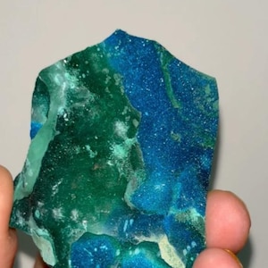 May include: A raw, irregular mineral specimen featuring a vibrant color palette of deep cobalt blue and forest green. The surface displays a crystalline, granular texture with natural variations in color intensity and light-reflecting facets.