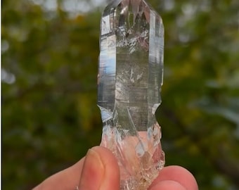 May include: A clear quartz crystal point with a faceted top and a rough, irregular base. The crystal is held up against a blurred green background, showcasing its transparency and natural form. The crystal is approximately 4 inches in length.