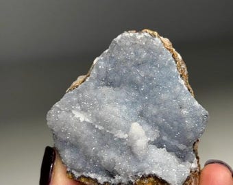 Puede incluir: Un grupo de cristales de cuarzo drusa de color azul claro con una matriz rugosa, marrón y dorada. El cristal tiene una textura brillante y granular y una pequeña formación de cristal blanco.