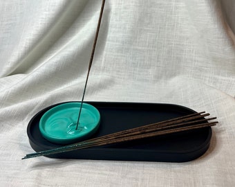 May include: A black oval incense holder with a teal ceramic dish in the centre. A single incense stick is standing upright in the dish, and several more incense sticks are lying on the holder.