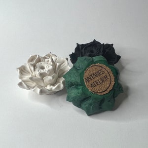 Puede incluir: Tres esculturas de flores de loto de cer&aacute;mica en blanco, negro y verde. La escultura verde tiene una etiqueta de corcho que dice "ANTARES ATELIER". Las flores de loto est&aacute;n detalladas con p&eacute;talos texturizados y una c&aacute;psula central.