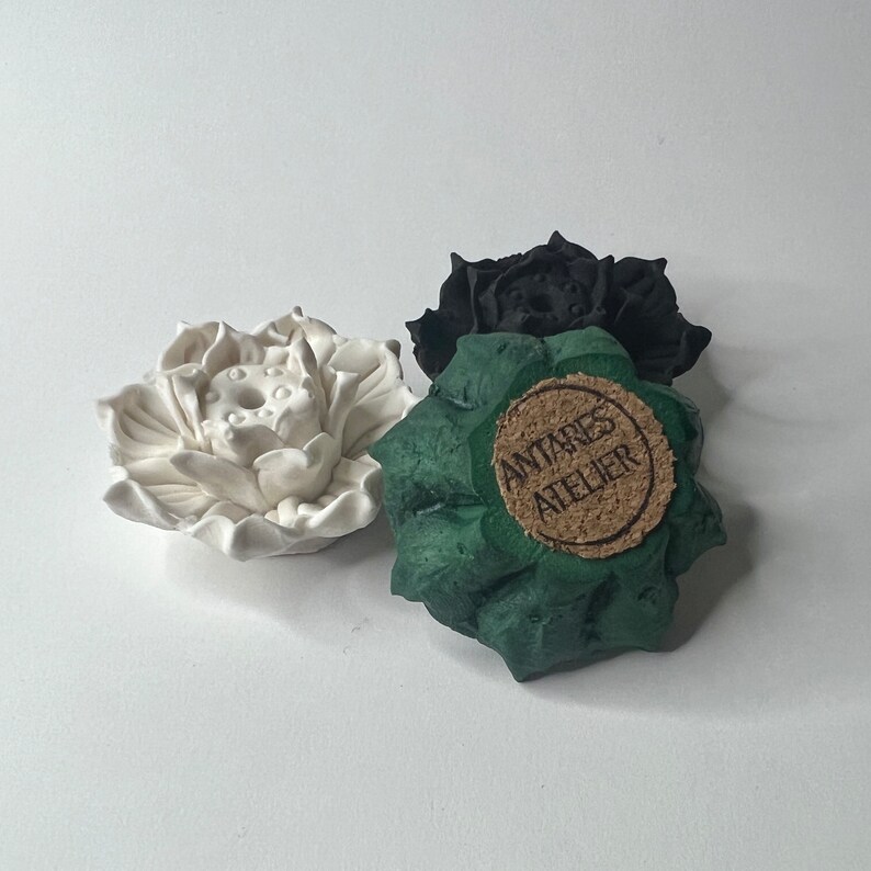 Puede incluir: Tres esculturas de flores de loto de cer&aacute;mica en blanco, negro y verde. La escultura verde tiene una etiqueta de corcho que dice "ANTARES ATELIER". Las flores de loto est&aacute;n detalladas con p&eacute;talos texturizados y una c&aacute;psula central.