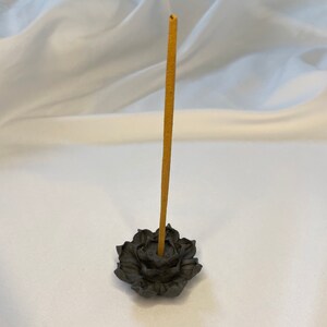 Puede incluir: Un porta incienso en forma de flor de loto gris oscuro con un solo palito de incienso marr&oacute;n. El porta incienso est&aacute; hecho de un material texturizado y se encuentra sobre una superficie blanca. El palito de incienso est&aacute; en posici&oacute;n vertical.
