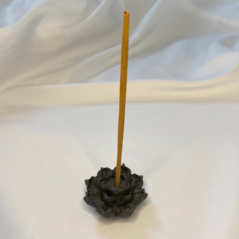 Puede incluir: Un porta incienso en forma de flor de loto gris oscuro con un solo palito de incienso marr&oacute;n. El porta incienso est&aacute; hecho de un material texturizado y se encuentra sobre una superficie blanca. El palito de incienso est&aacute; en posici&oacute;n vertical.
