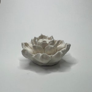Puede incluir: Un portavelas de cer&aacute;mica blanca con forma de flor de loto. El portavelas tiene forma de flor de loto con m&uacute;ltiples p&eacute;talos. Los p&eacute;talos son detallados y texturizados. El portavelas est&aacute; sobre una superficie blanca.