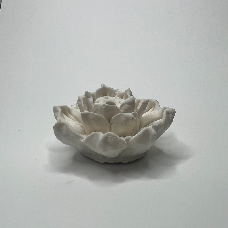 Puede incluir: Un portavelas de cer&aacute;mica blanca con forma de flor de loto. El portavelas tiene forma de flor de loto con m&uacute;ltiples p&eacute;talos. Los p&eacute;talos son detallados y texturizados. El portavelas est&aacute; sobre una superficie blanca.