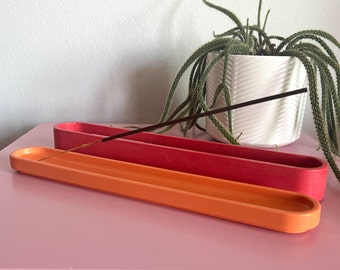 Puede incluir: Dos porta inciensos de cerámica, uno naranja y otro rojo, con un palo de incienso marrón descansando sobre el porta incienso naranja. Los porta inciensos están sobre una superficie rosa.