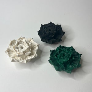Puede incluir: Tres esculturas decorativas de flores de loto en blanco, negro y verde. Las esculturas tienen dise&ntilde;os detallados de p&eacute;talos y una c&aacute;psula central de semillas. Los objetos est&aacute;n dispuestos sobre una superficie blanca, mostrando el contraste de colores y texturas.