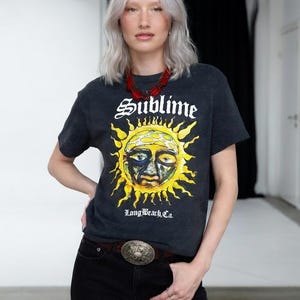 May include: Dark gray t-shirt with the word "Sublime" above a yellow and orange sun graphic. The text "Long Beach, Ca." is below the sun. The person is wearing black jeans and a brown belt with a silver buckle.