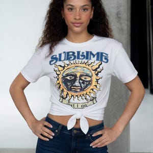 May include: White t-shirt with a sun graphic and the word "SUBLIME" in blue. The sun graphic is yellow and black with a face in the center. The shirt is knotted at the waist and paired with dark blue jeans.