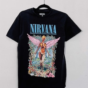 Puede incluir: Una camiseta negra de manga corta con la palabra NIRVANA en letras azul claro. El gráfico muestra una figura alada con anatomía expuesta en un bosque rodeada de flores, enredaderas y un pequeño reloj, todo en colores vibrantes.