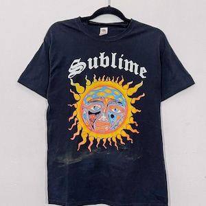 以下が含まれることがあります： 黒の半袖Tシャツ。白いゴシック体で「Sublime」という文字がプリントされ、その下に多色使いの顔、青とオレンジの光線、そして青、オレンジ、赤の複雑な模様が描かれた、様式化された太陽のグラフィックが配置されています。