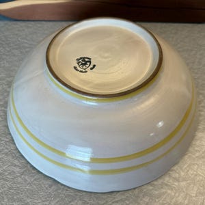 May include: A handmade ceramic bowl with a white glaze and two yellow stripes. The bowl has a rounded shape and a brown rim. The bottom of the bowl has a stamp. This bowl is perfect for serving soups, salads, or other dishes.