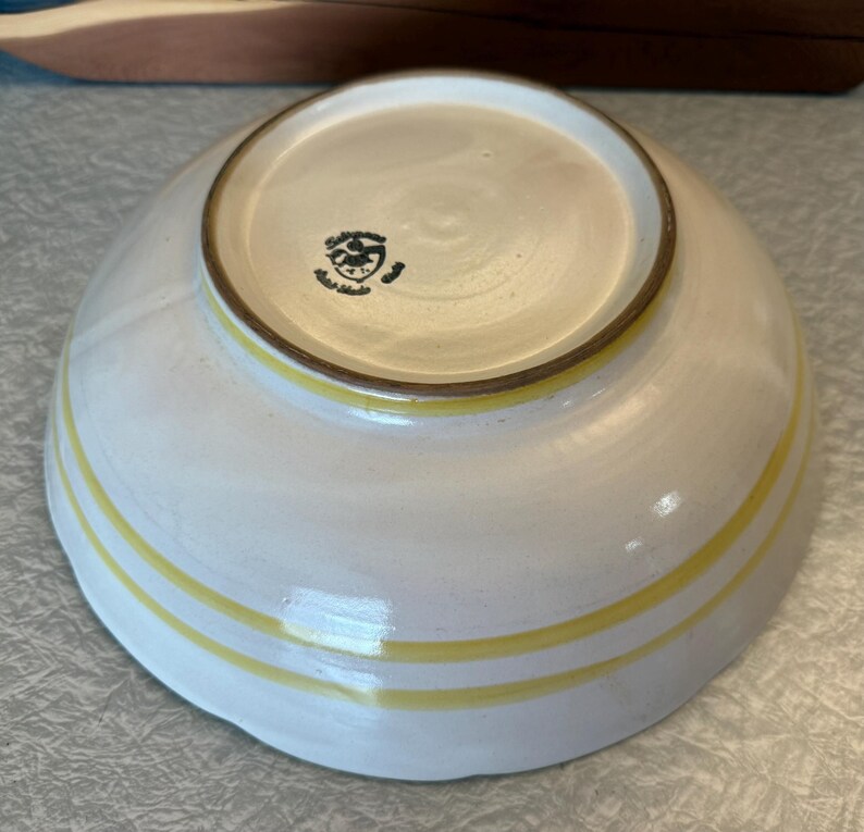 May include: A handmade ceramic bowl with a white glaze and two yellow stripes. The bowl has a rounded shape and a brown rim. The bottom of the bowl has a stamp. This bowl is perfect for serving soups, salads, or other dishes.