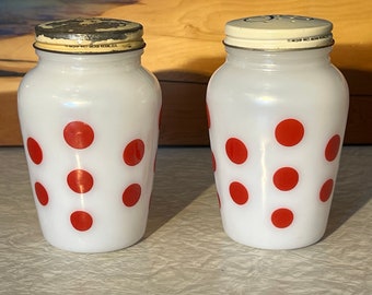 Op de afbeelding: Twee vintage zout- en peperstrooiers van wit glas met rode stippen. Elke strooier heeft een metalen deksel met kleine gaatjes om te doseren. De strooiers zijn cilindrisch met een licht uitlopende basis.