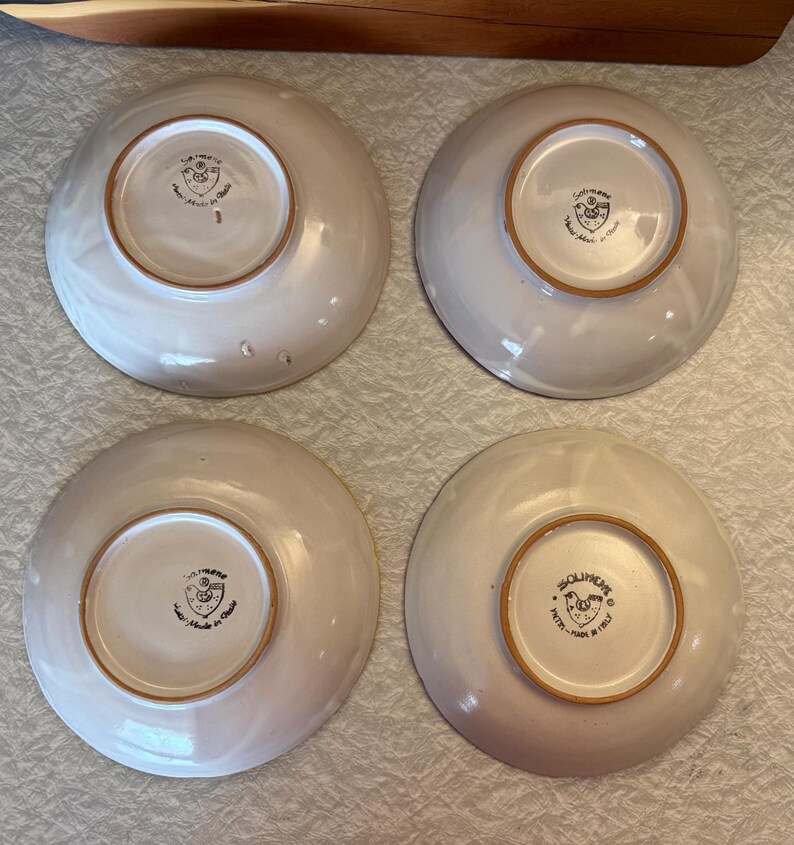 May include: Four off-white ceramic bowls with a slightly textured surface. Each bowl has a brown rim and a central design with text. The bowls are arranged in a square formation, showcasing their underside.