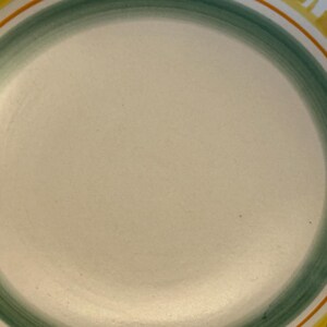 May include: A round, off-white ceramic plate with a green and orange border. The plate has a yellow rim with a leaf design. The plate is suitable for serving food.