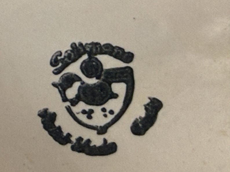 May include: A black, stylized emblem with the word "Collingwood" arched above a central design. The design includes a shield shape with a bird-like figure and several small dots. The emblem is on a cream-colored background.