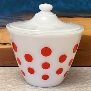 May include: White glass jar with a lid, decorated with large red polka dots. The jar has a rounded shape and a matching white lid with a small knob. A vintage-style kitchen storage container.