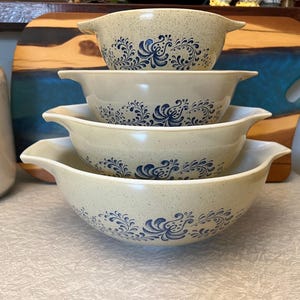 Peut inclure: Une pile de quatre saladiers beiges avec des motifs floraux bleus. Chaque bol a une poignée de chaque côté. Les bols sont disposés par ordre de taille décroissante, le plus grand bol étant en bas.