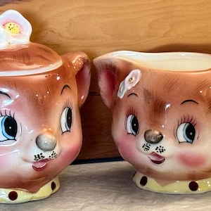 May include: A ceramic sugar bowl with a lid and a matching creamer, both shaped like brown bear faces with pink cheeks, blue eyes, and yellow bases featuring brown polka dots. The sugar bowl lid is topped with two small white and blue flowers.