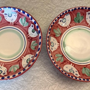 May include: Two hand-painted ceramic plates with a red background and a white, green, and blue design. The plates feature a central white circle, surrounded by a green and blue ring. The outer rim has a blue dotted border and a repeating pattern of white birds and green leaves.