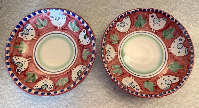 May include: Two hand-painted ceramic plates with a red background and a white, green, and blue design. The plates feature a central white circle, surrounded by a green and blue ring. The outer rim has a blue dotted border and a repeating pattern of white birds and green leaves.