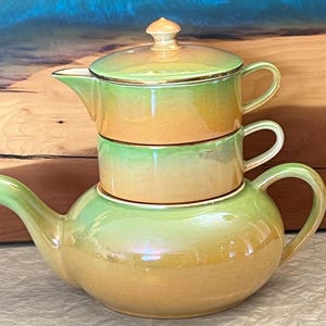 May include: A three-piece tea set with an iridescent finish. The set comprises a teapot, a teacup, and a lidded creamer. The colours shift from light green to golden yellow, with gold trim. The teapot has a curved spout and a rounded body.