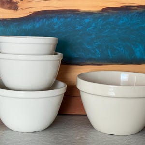 May include: A set of four off-white ceramic mixing bowls of varying sizes. Three bowls are stacked vertically in a column, while the fourth bowl sits independently to the right. Each bowl features a smooth, rounded design with a thick rim.