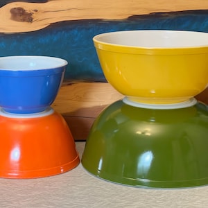 May include: Two sets of vintage mixing bowls in bright colors. The smaller set features a blue bowl atop an orange bowl. The larger set has a yellow bowl on a green bowl. The bowls have white interiors.
